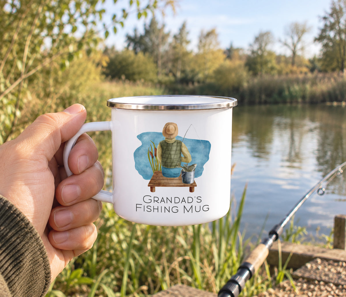 A hand holds a white enamel-style mug with a silver rim in front of an outdoor lakeside scene. The mug has a printed illustration of a person fishing and the text 'GRANDAD'S FISHING MUG'.