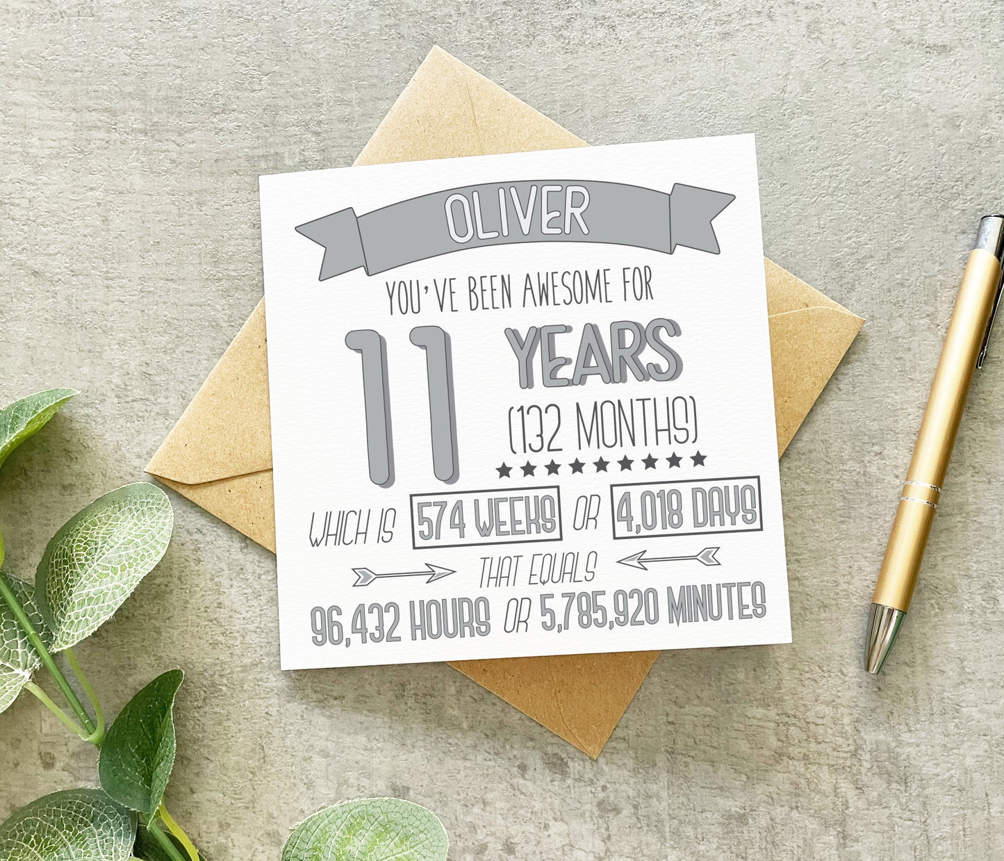 A personalized 11th birthday card with text that calculates and displays the time in years, months, weeks, days, hours, and minute in grey, placed on a table with a pen and green leaves in the background.