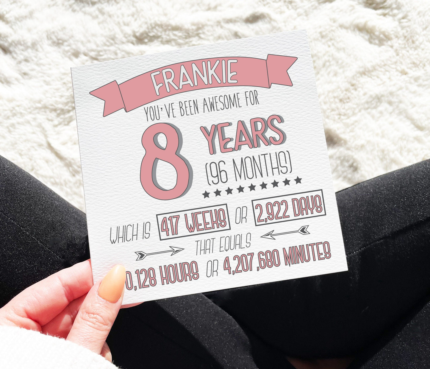 A pink personalised 8th birthday card with text that calculates and prints the time in years, months, weeks, days, hours, and minutes, placed on a table with a pen and some greenery in the background.