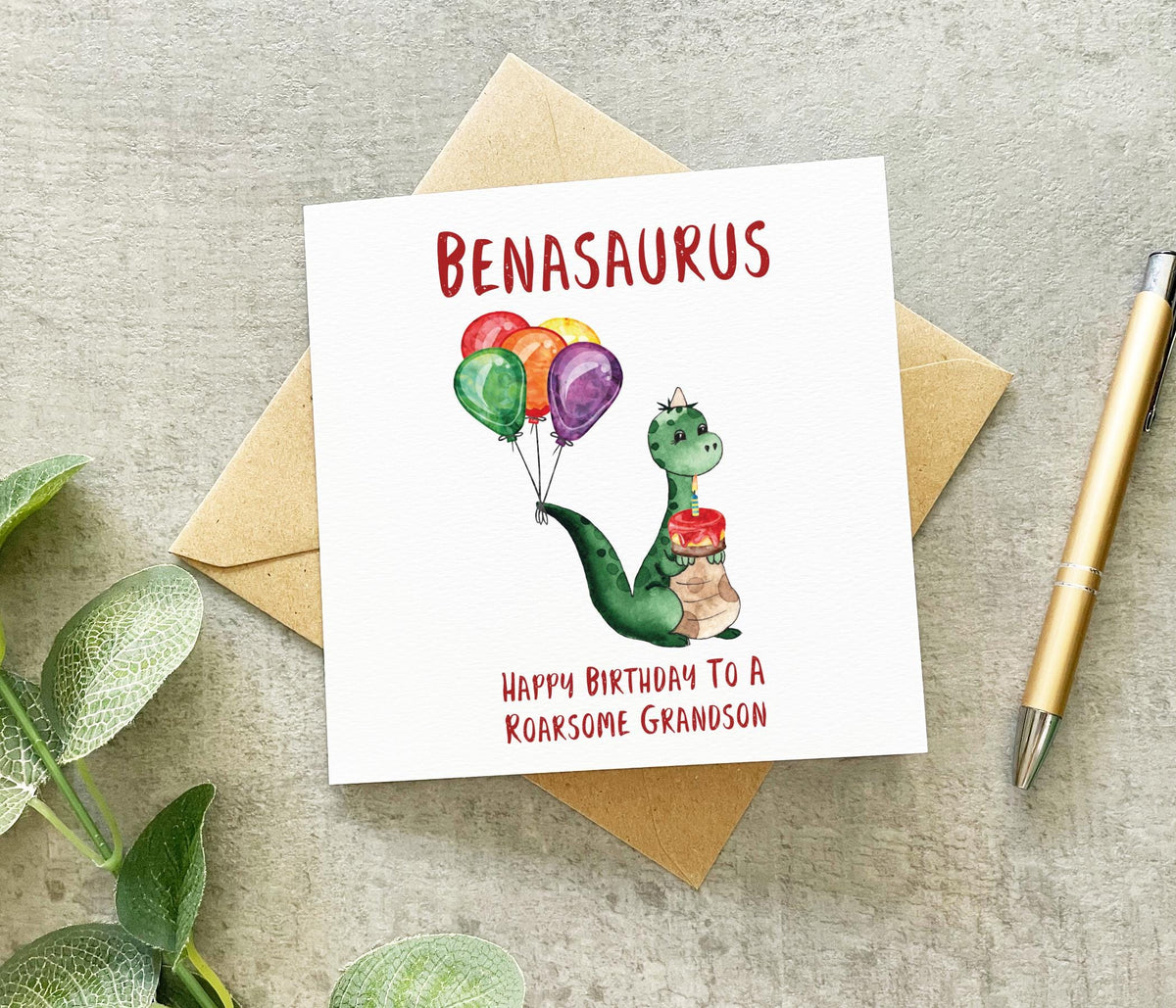 Greeting card with an illustration of a dinosaur on it, with 'Happy Birthday to a roarsome grandson' text and the name 'Bensaurus' (or a similar spelling). The card is presented on a table with a gold pen and green leaves in the background.