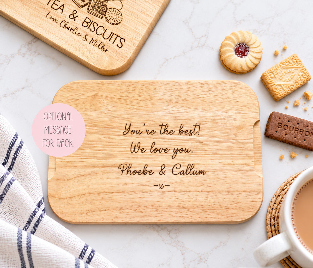 A personalised wooden tea and biscuits board showing the back of the board engraved with 'You're the best! We love you. Phoebe & Callum -u-' is displayed on a white marble surface with tea time props including biscuits, a striped towel, and a cup of tea.