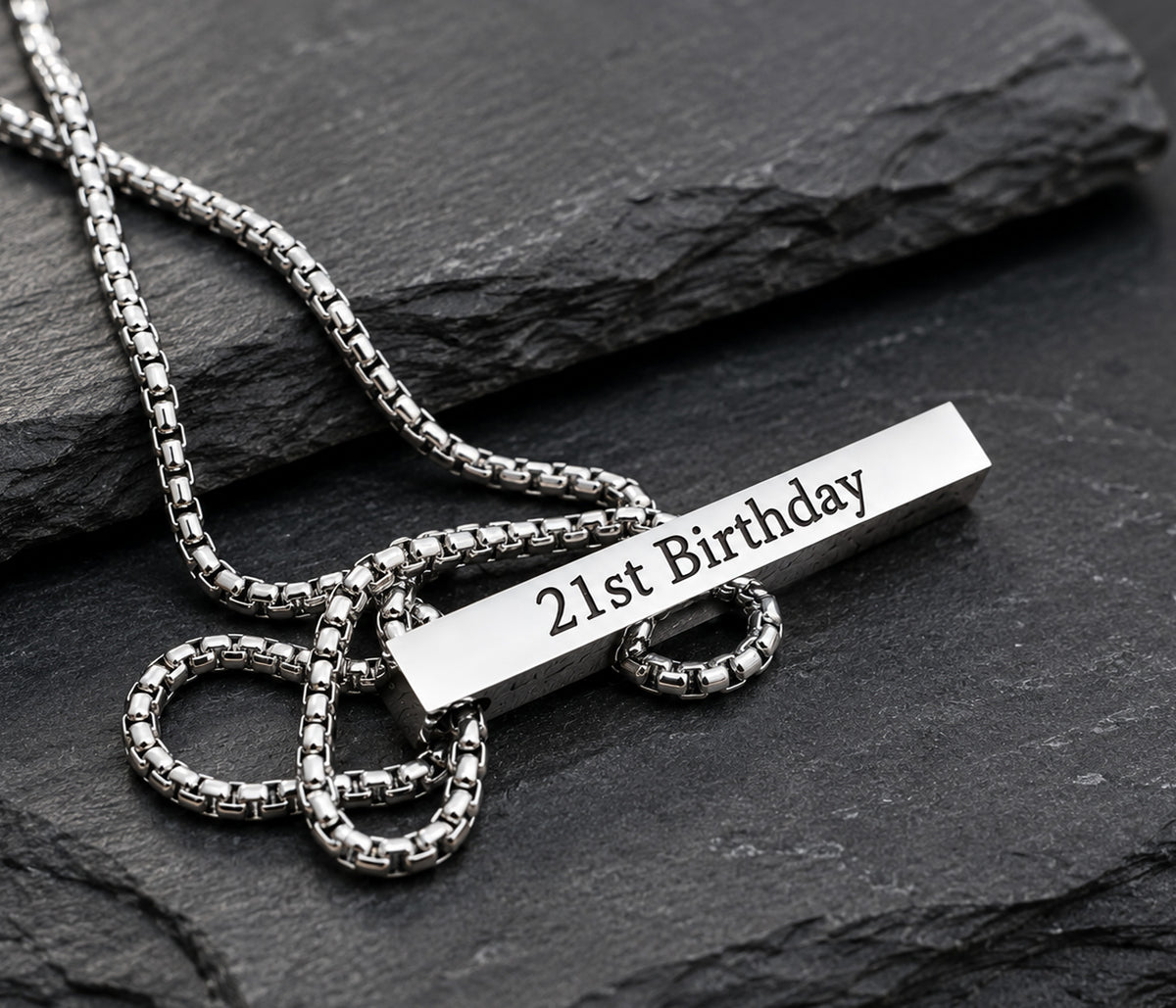 A silver-toned metal necklace with a box chain and a rectangular bar pendant engraved with '21st Birthday', displayed on a dark slate surface.