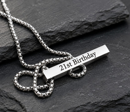 A silver-toned metal necklace with a box chain and a rectangular bar pendant engraved with '21st Birthday', displayed on a dark slate surface.