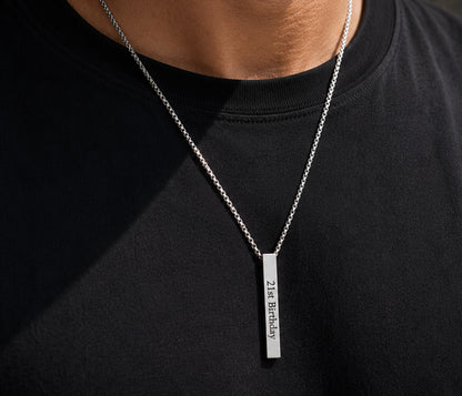 A close-up of a person wearing a silver chain necklace with a vertical rectangular bar engraved with '21st Birthday' on a black t-shirt.