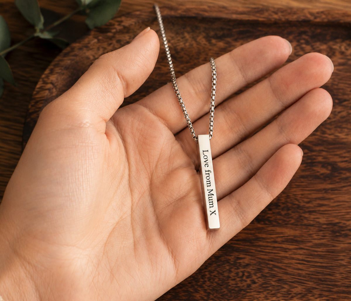 A silver bar necklace engraved with 'Love from Mom X' resting on an open hand over a dark wooden surface.