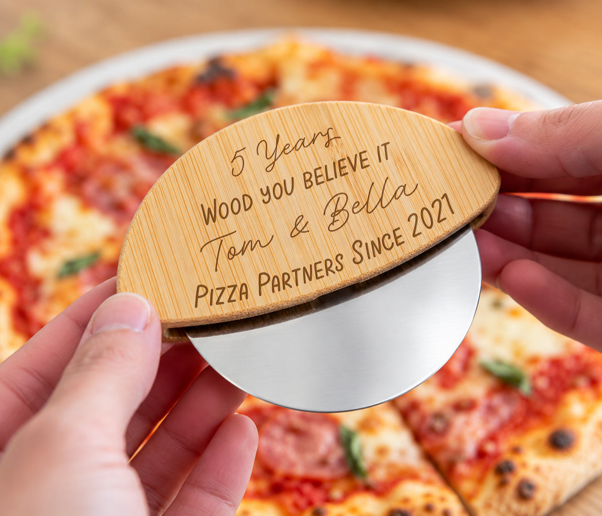 Wooden handle pizza wheel held over a pizza with the wording engraved - 5 years, would you believe it, two names and then 'Pizza partners since 2021