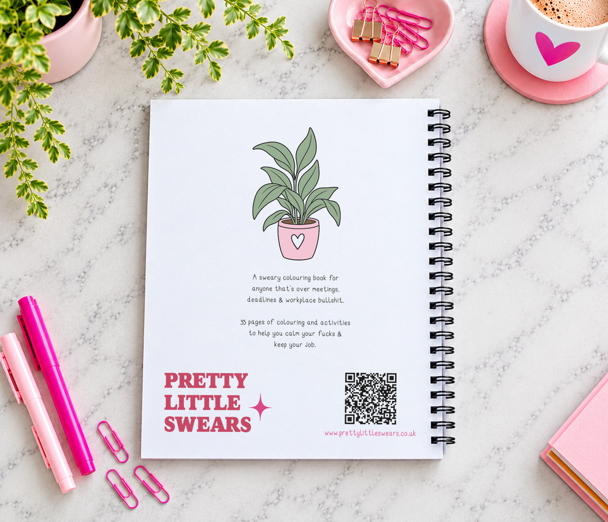 A spiral-bound notebook titled 'PRETTY LITTLE SWEARS' lies on a light marble surface, surrounded by pink stationery items, a plant, and a cup of coffee. The notebook cover features a simple illustration of a potted plant and product text.