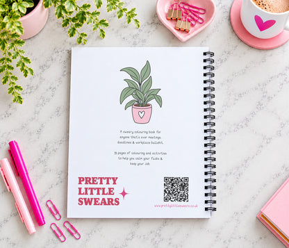 A spiral-bound notebook titled 'PRETTY LITTLE SWEARS' lies on a light marble surface, surrounded by pink stationery items, a plant, and a cup of coffee. The notebook cover features a simple illustration of a potted plant and product text.