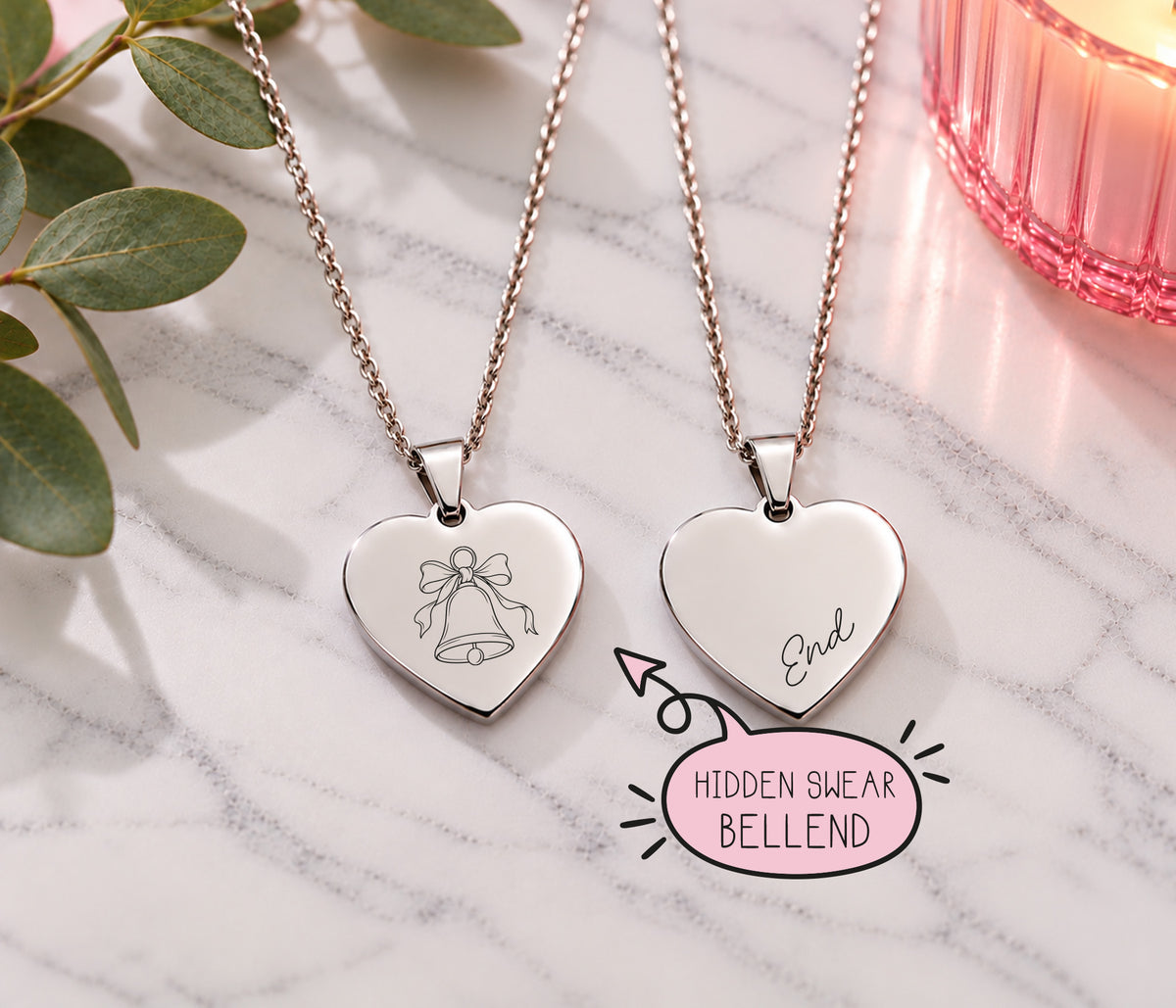 Two silver heart pendant necklaces on chains, displayed on a white marble surface with green leaves and a pink candle. One heart is engraved with a bell and bow, and the other with 'End'.