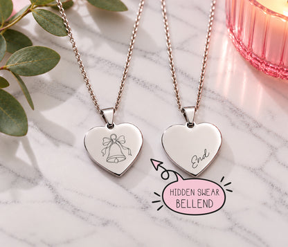 Two silver heart pendant necklaces on chains, displayed on a white marble surface with green leaves and a pink candle. One heart is engraved with a bell and bow, and the other with 'End'.