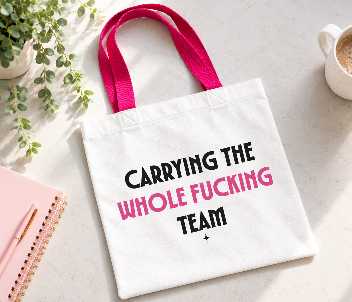 A white canvas tote bag with bright pink handles, featuring the printed text 'CARRYING THE WHOLE FUCKING TEAM' in black and pink lettering. The bag is displayed in a styled flat-lay setting on a light gray surface, surrounded by a pink notebook, a small potted plant, and a white coffee mug.
