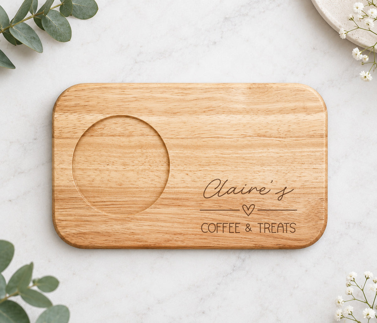 A rectangular wooden serving board with rounded corners, featuring a circular recessed area on the left and engraved text reading 'Claire's COFFEE & TREATS' with a small heart. The board is photographed from above on a light marble surface with decorative greenery and small white flowers around it.