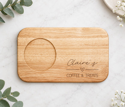 A rectangular wooden serving board with rounded corners, featuring a circular recessed area on the left and engraved text reading 'Claire's COFFEE & TREATS' with a small heart. The board is photographed from above on a light marble surface with decorative greenery and small white flowers around it.