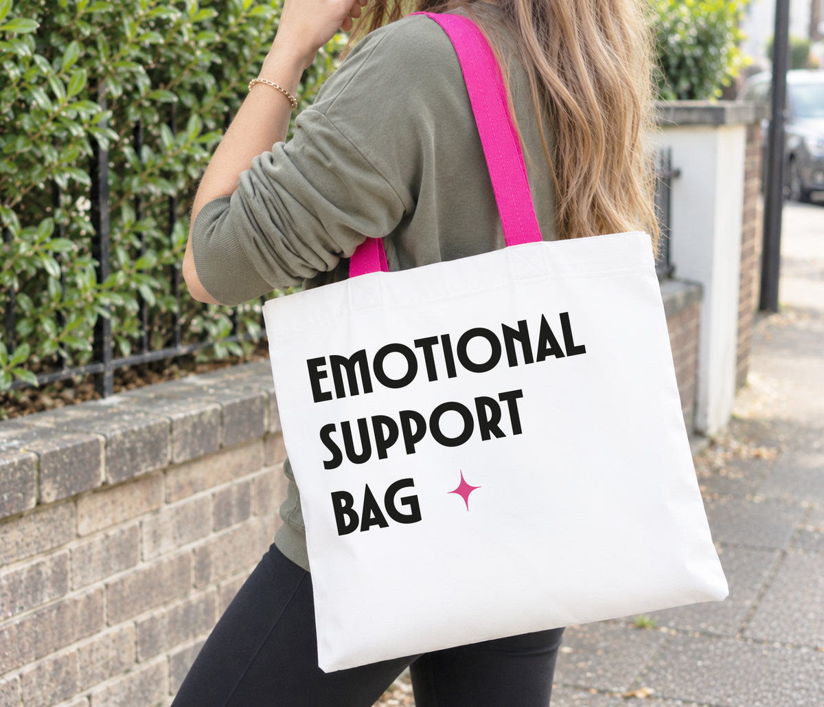Large white tote bag with bright pink handles. The bag is printed with big bold black text saying emotional support bag and a pink sparkle.
