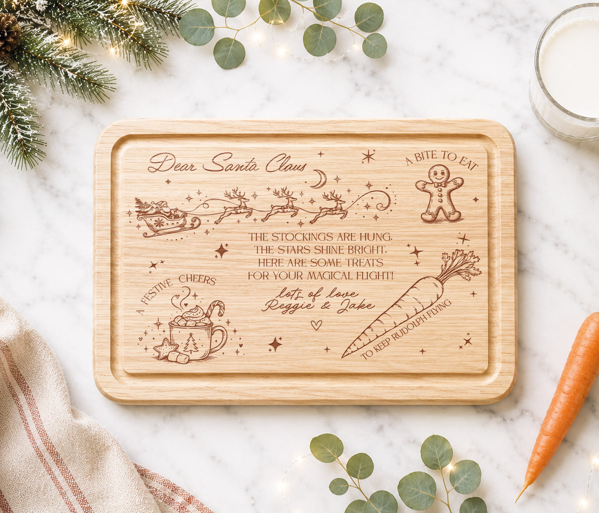 A rectangular wooden rectangular christmas eve plate with rounded corners, featuring engraved Christmas-themed illustrations and a personalized holiday message. The board is styled in a flat-lay on a white marble surface with eucalyptus leaves, a carrot, a glass of milk, and a striped napkin.