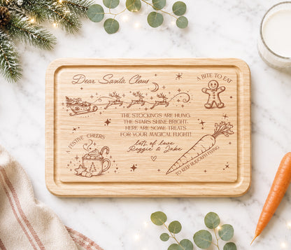 A rectangular wooden rectangular christmas eve plate with rounded corners, featuring engraved Christmas-themed illustrations and a personalized holiday message. The board is styled in a flat-lay on a white marble surface with eucalyptus leaves, a carrot, a glass of milk, and a striped napkin.