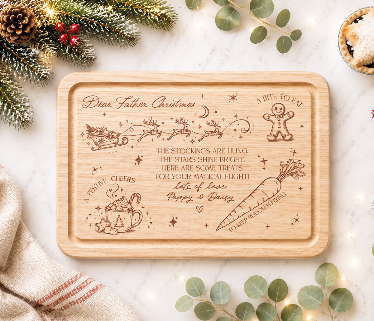 A rectangular wooden Christmas eve board with rounded corners, featuring engraved festive illustrations and the text 'Dear Father Christmas' and holiday-themed phrases. The tray is photographed in a styled flat-lay setting with pine branches, eucalyptus leaves, a napkin, and a small pie on a light marble surface.