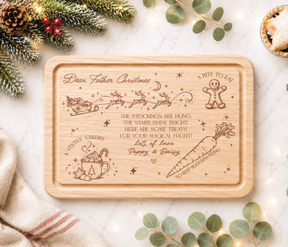 A rectangular wooden Christmas eve board with rounded corners, featuring engraved festive illustrations and the text 'Dear Father Christmas' and holiday-themed phrases. The tray is photographed in a styled flat-lay setting with pine branches, eucalyptus leaves, a napkin, and a small pie on a light marble surface.