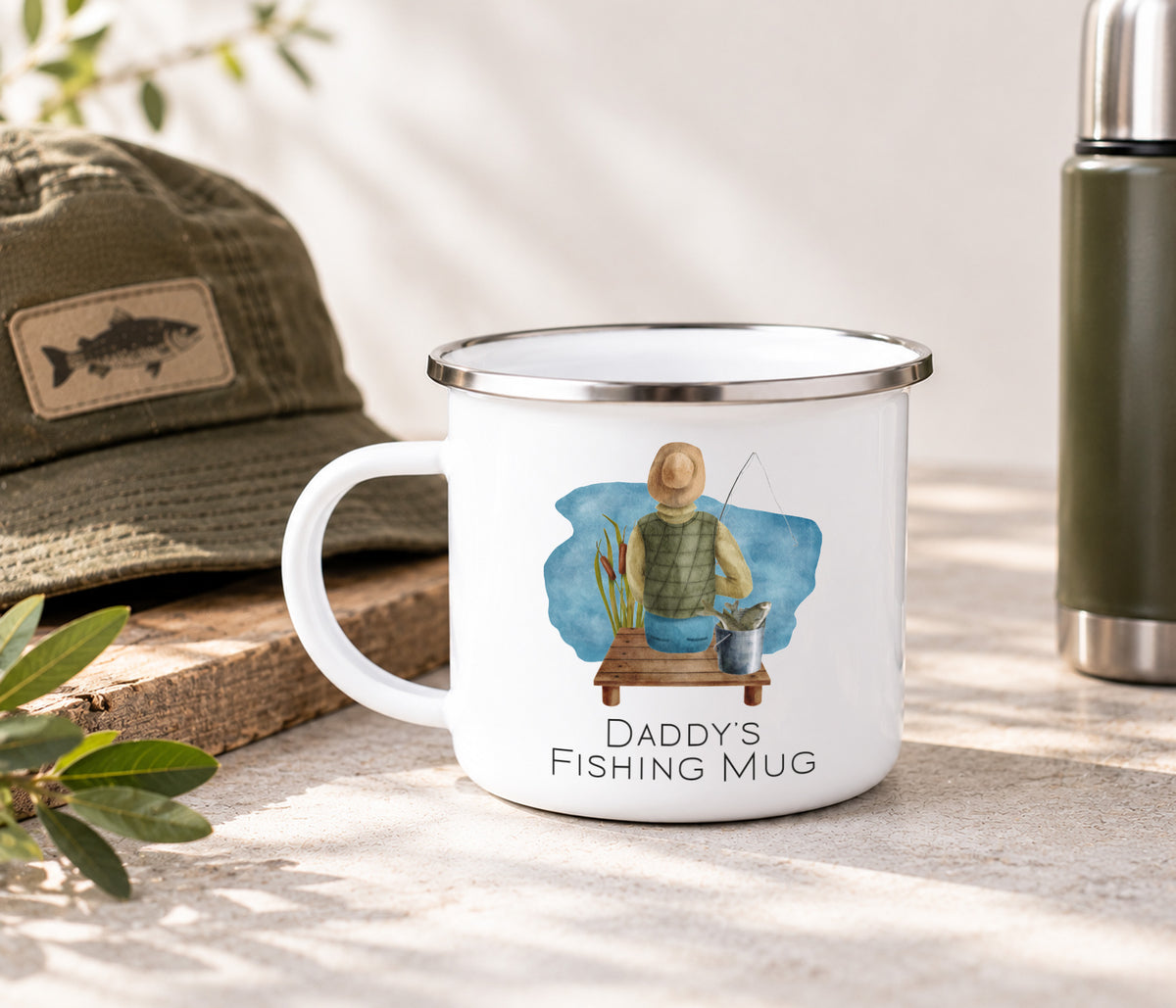 A white enamel-style mug with a silver rim and handle, printed with an illustration of a man fishing and the text 'Daddy's Fishing Mug.' The mug is styled outdoors on a stone surface with a green cap, leaves, and a green bottle in the background.