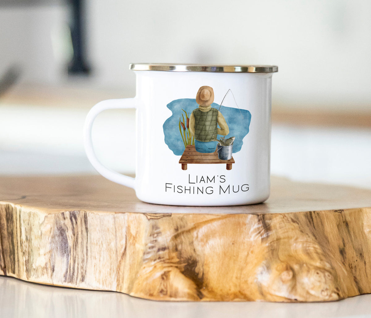A white enamel-style mug with a silver rim sits on a rustic wood slice. The mug features a watercolor illustration of a man fishing and the text 'Liam's Fishing Mug'.