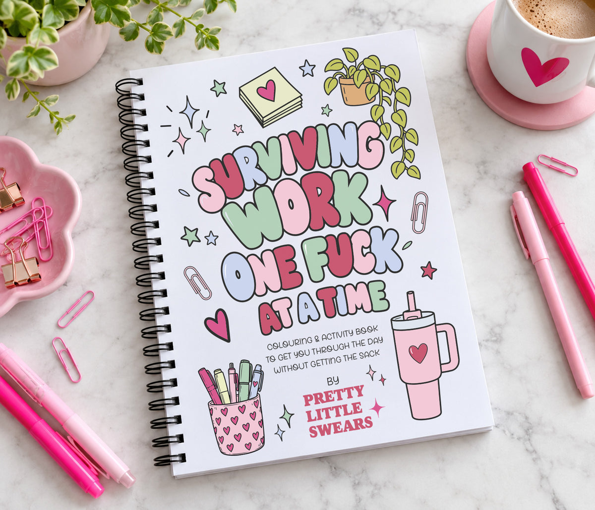 A spiral-bound notebook with a white cover, featuring colorful hand-drawn text that reads 'Surviving Work One Fuck At A Time' and cute illustrations, styled on a marble surface with pink pens, paperclips, and a coffee cup.