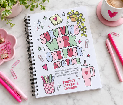 A spiral-bound notebook with a white cover, featuring colorful hand-drawn text that reads 'Surviving Work One Fuck At A Time' and cute illustrations, styled on a marble surface with pink pens, paperclips, and a coffee cup.