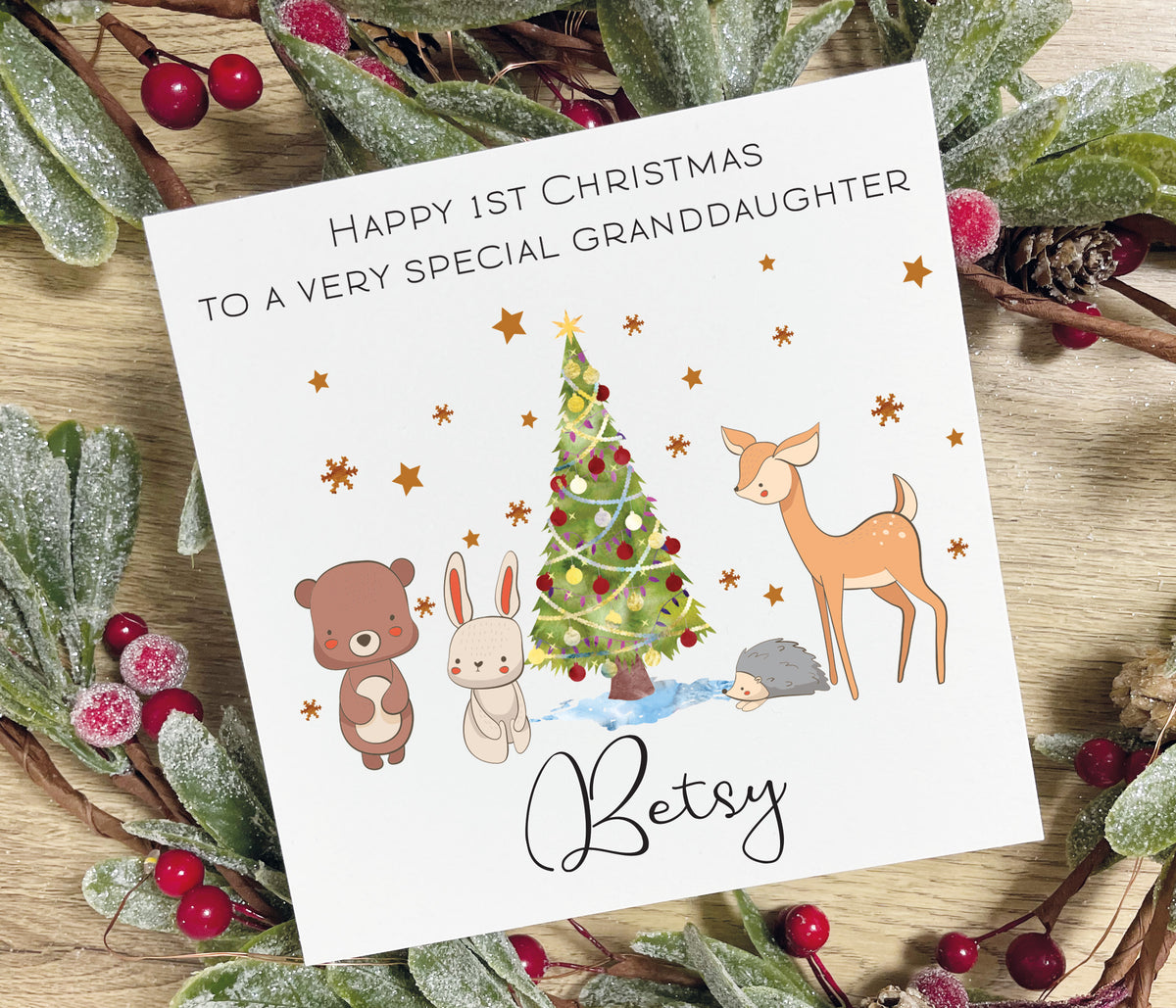 A personalized Christmas card for a 1st Christmas Granddaughter with woodland animal illustrations, styled on a rustic wooden surface with a wreath of mistletoe and red berries. The card is addressed to 'Betsy' and features a tree, bear, rabbit, deer, and hedgehog.
