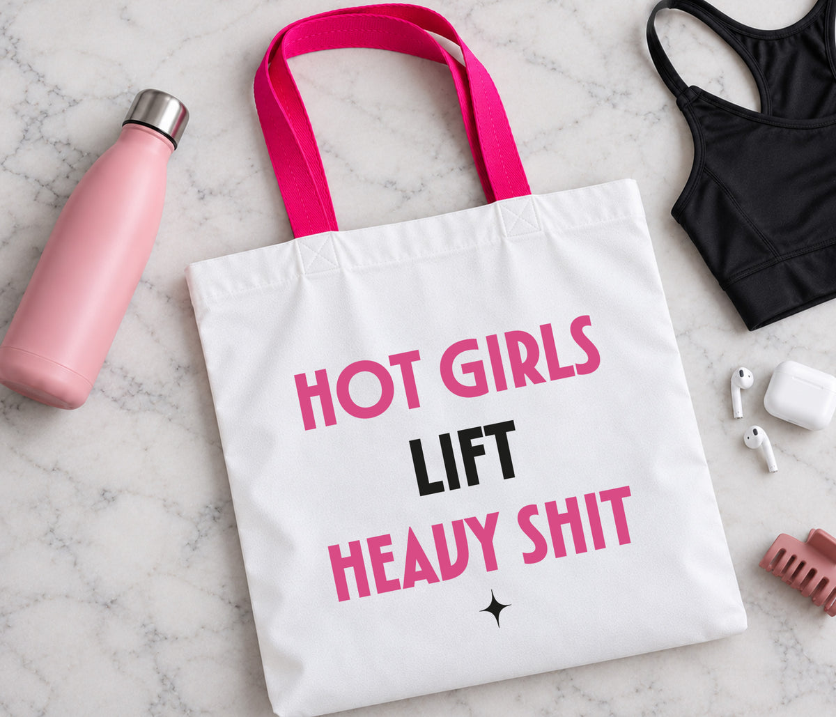 A white canvas gym tote bag with hot pink and black text reading 'HOT GIRLS LIFT HEAVY SHIT' is displayed in a flat-lay arrangement on a white marble surface, surrounded by lifestyle props including a pink water bottle, a black sports bra, earbuds, and a hair clip.