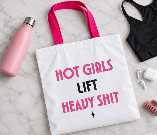 A white canvas gym tote bag with hot pink and black text reading 'HOT GIRLS LIFT HEAVY SHIT' is displayed in a flat-lay arrangement on a white marble surface, surrounded by lifestyle props including a pink water bottle, a black sports bra, earbuds, and a hair clip.
