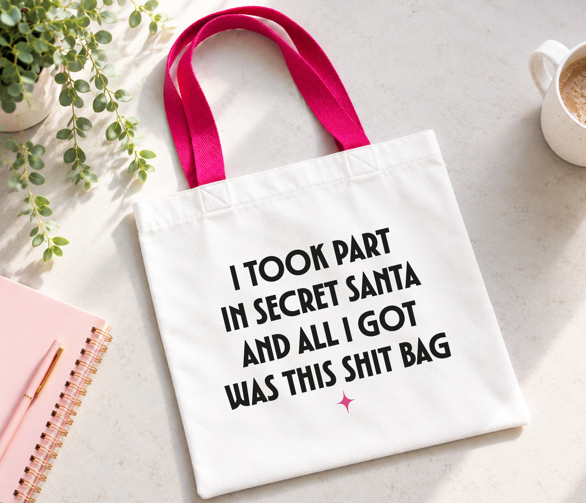 A white canvas tote bag with bright pink handles, featuring the humorous text 'I TOOK PART IN SECRET SANTA AND ALL I GOT WAS THIS SHIT BAG' in black lettering. The bag is displayed in a styled flat-lay setting on a light gray surface, surrounded by a pink notebook, a small potted plant, and a white coffee mug.