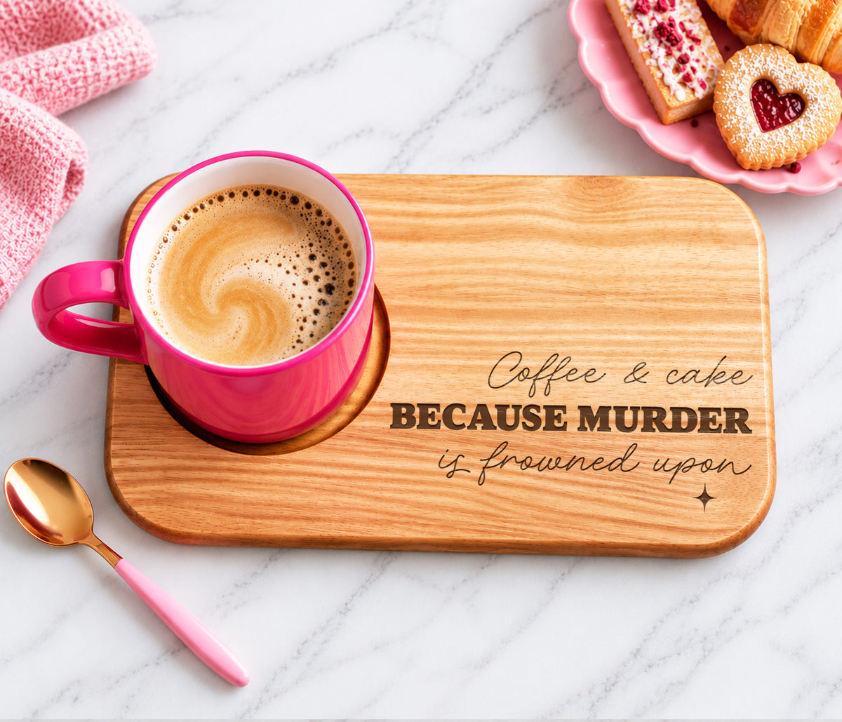 A wooden coffee and cake biscuit tray engraved with the phrase 'Coffee & cake BECAUSE MURDER is frowned upon', styled with a pink mug of coffee, a pink spoon, and pastries on a marble surface.