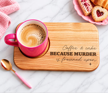 A wooden coffee and cake biscuit tray engraved with the phrase 'Coffee & cake BECAUSE MURDER is frowned upon', styled with a pink mug of coffee, a pink spoon, and pastries on a marble surface.