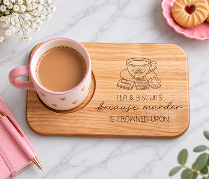 A rectangular wooden tray with rounded corners sits on a white marble surface. The tray is engraved with a teacup, biscuits, and the phrase 'TEA & BISCUITS because murder IS FROWNED UPON.