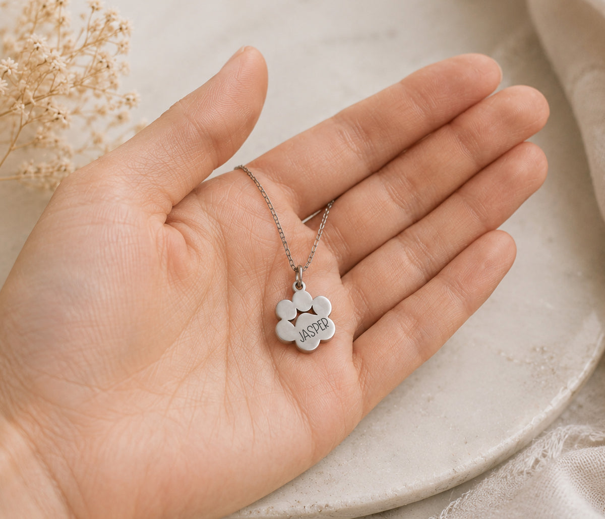 A silver paw print shaped pendant necklace engraved with the name 'JASPER' is displayed on an open hand.