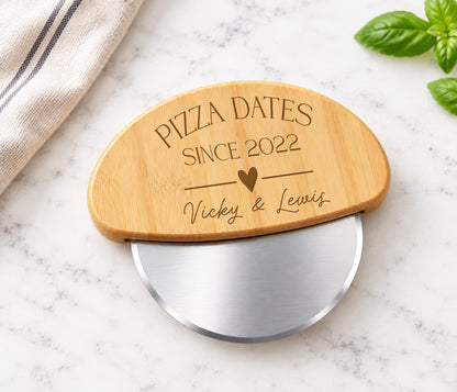 A personalized wooden pizza cutter with a brushed silver metal blade, engraved with 'PIZZA DATES SINCE 2022' and 'Vicky & Lewis' with a heart. The product is styled on a white marble surface with a striped kitchen towel and fresh basil leaves.