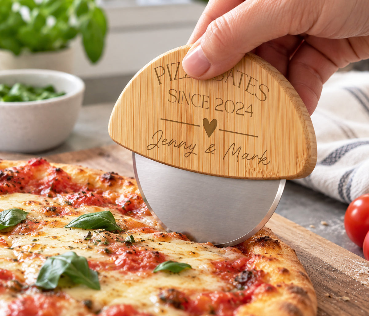 A hand holds a wooden pizza cutter with a brushed silver metal wheel, engraved with 'PIZZA DATES SINCE 2024' and 'Jenny & Mark' with a heart. The cutter is slicing a freshly baked pepperoni and cheese pizza on a wooden board, with a bowl of basil and a striped kitchen towel in the background.