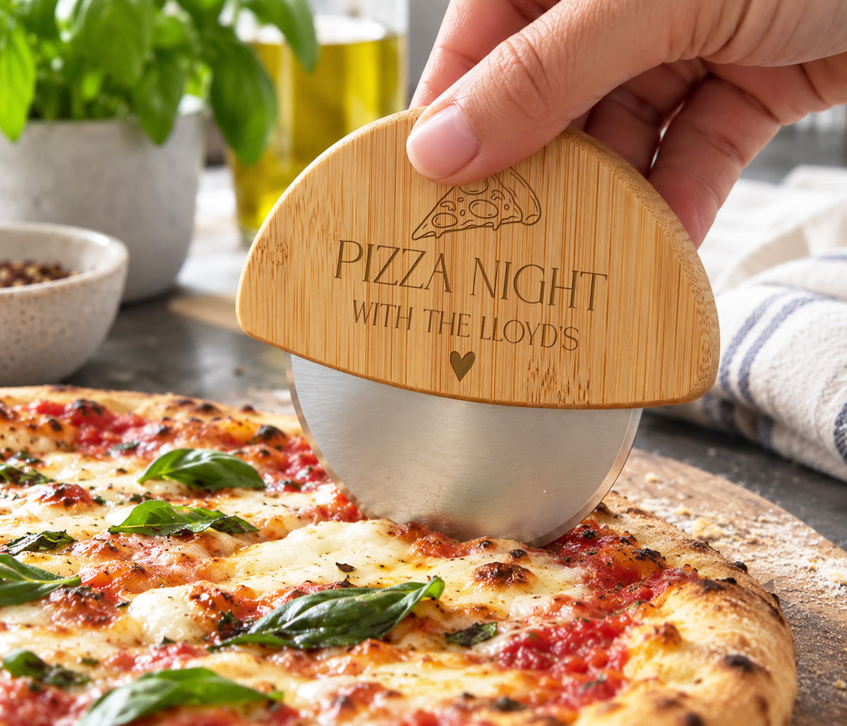 A hand holding a bamboo pizza cutter with the engraved text 'PIZZA NIGHT WITH THE LLOYDS' over a freshly baked pizza on a wooden board.