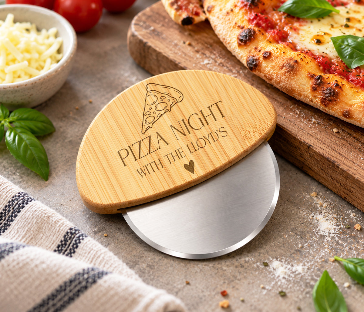 A wooden pizza cutter with a brushed silver metal wheel, featuring an engraved bamboo lid that reads 'PIZZA NIGHT WITH THE LLOYDS' and shows a pizza slice graphic. The product is styled on a rustic stone surface with a pizza, basil, cheese, and a striped towel.