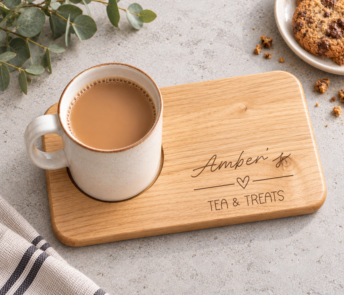 A rectangular wooden tea and treats tray engraved with 'Amber's TEA & TREATS' and a heart, with an indent holding a ceramic mug filled with tea. The tray is styled on a light gray surface with a cookie, crumbs, eucalyptus leaves, and a striped cloth.