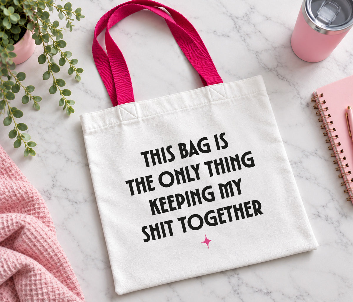 A white canvas tote bag with bright pink handles and black printed text reading 'THIS BAG IS THE ONLY THING KEEPING MY SHIT TOGETHER' on a light marble surface, styled with a pink towel, plant, notebook, and tumbler.