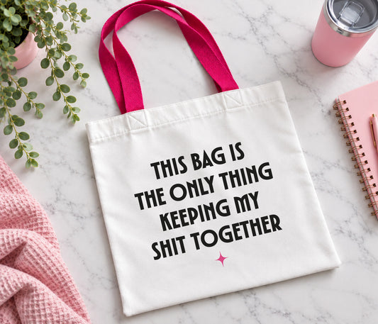 A white canvas tote bag with bright pink handles and black printed text reading 'THIS BAG IS THE ONLY THING KEEPING MY SHIT TOGETHER' on a light marble surface, styled with a pink towel, plant, notebook, and tumbler.
