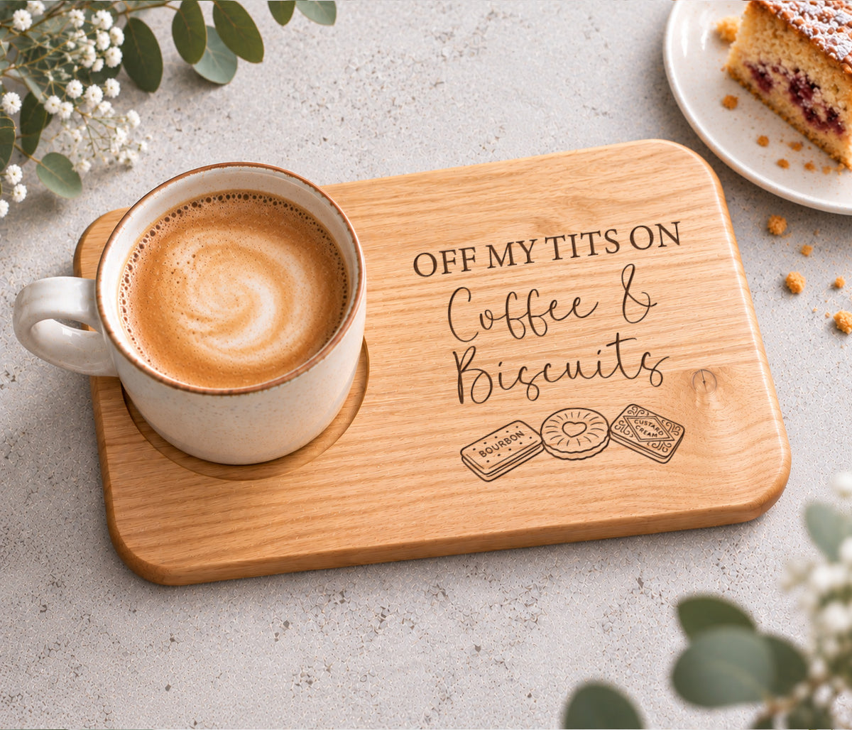 A rectangular wooden serving tray with rounded corners, engraved with the phrase 'OFF MY TITS ON Coffee & Biscuits' and small biscuit illustrations, styled with a cup of coffee on a light stone surface.