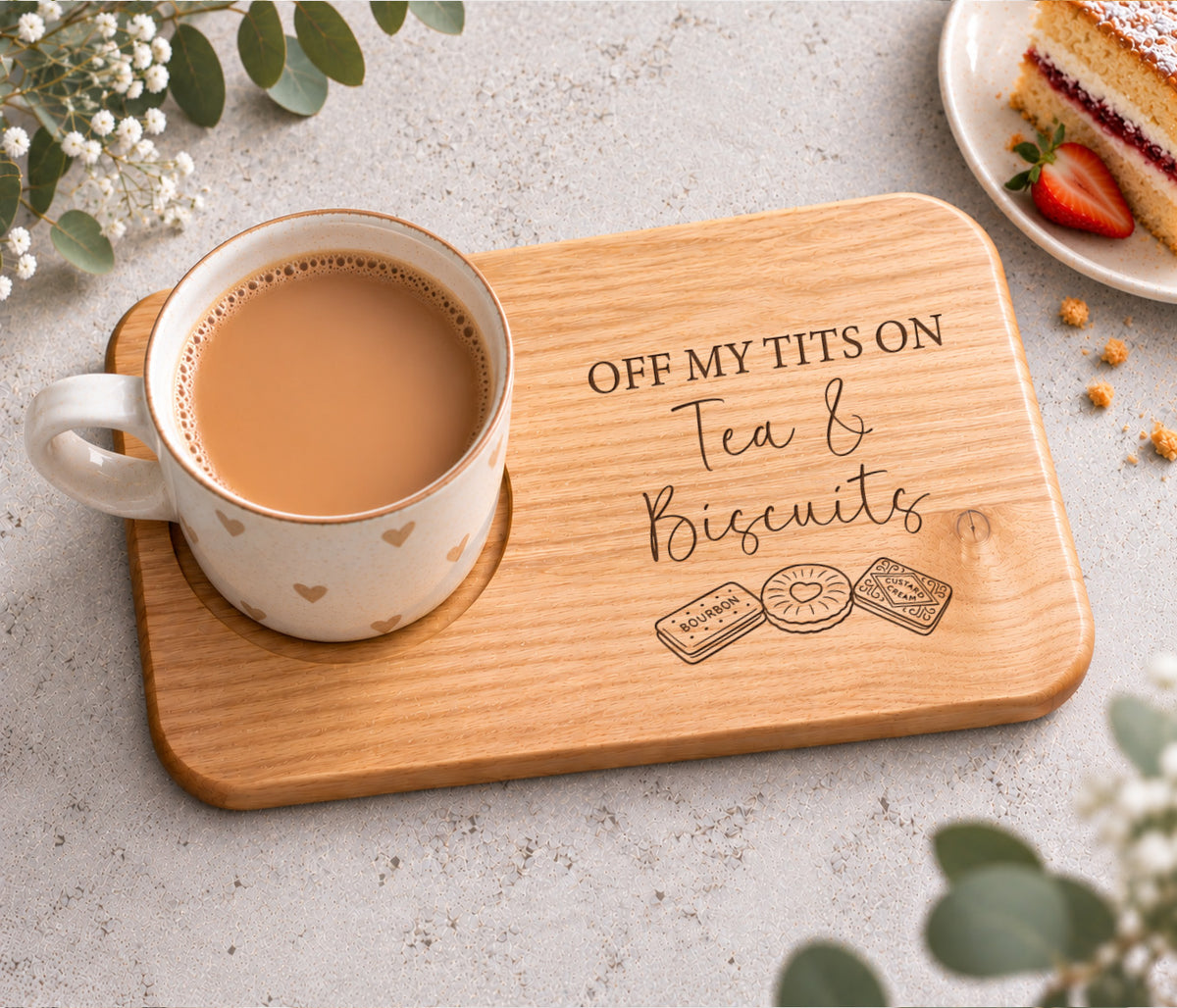 A rectangular wooden tea and biscuits tray engraved with the phrase 'OFF MY TITS ON Tea & Biscuits' and small biscuit illustrations, styled with a white heart-patterned mug filled with tea on a light stone surface, with decorative flowers and a slice of cake in the corners.